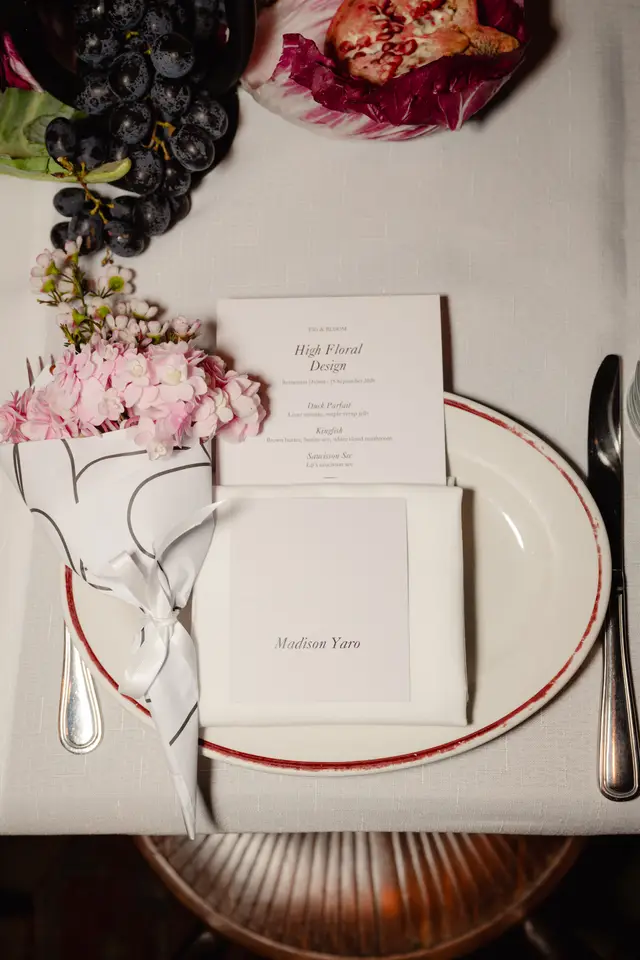 An overhead shot of an elegant place setting on a white linen tablecloth. The setting includes a white plate with a red rim, silverware, a folded white napkin, a menu card for 'High Floral Design' by 'Fig & Bloom', and a name card for 'Madison Yaro'. A small bouquet of pink hydrangeas and white wax flowers is wrapped in patterned paper, and a cluster of dark grapes, radicchio, and a half pomegranate are also visible.