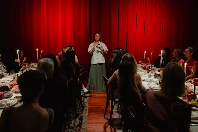 An elegant indoor event features a woman speaking to a group of guests seated at a long, candlelit table. A vibrant red curtain serves as the backdrop, creating a warm and intimate atmosphere. The table is adorned with floral arrangements, candles, and place settings.