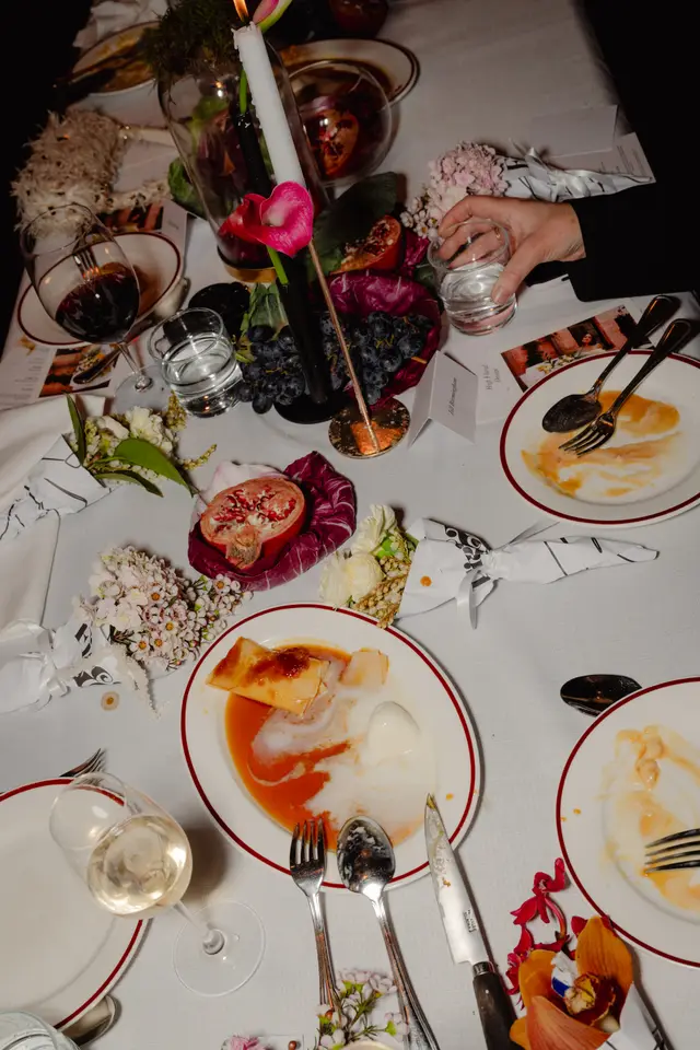 A top-down view of an elegantly set dinner table at an event, featuring partially eaten dishes, wine and water glasses, and a variety of floral arrangements and fruits. A lit white taper candle adds to the atmospheric lighting, and a hand is visible holding a water glass. The table is adorned with white linen, red-rimmed plates, and small wrapped floral favors.