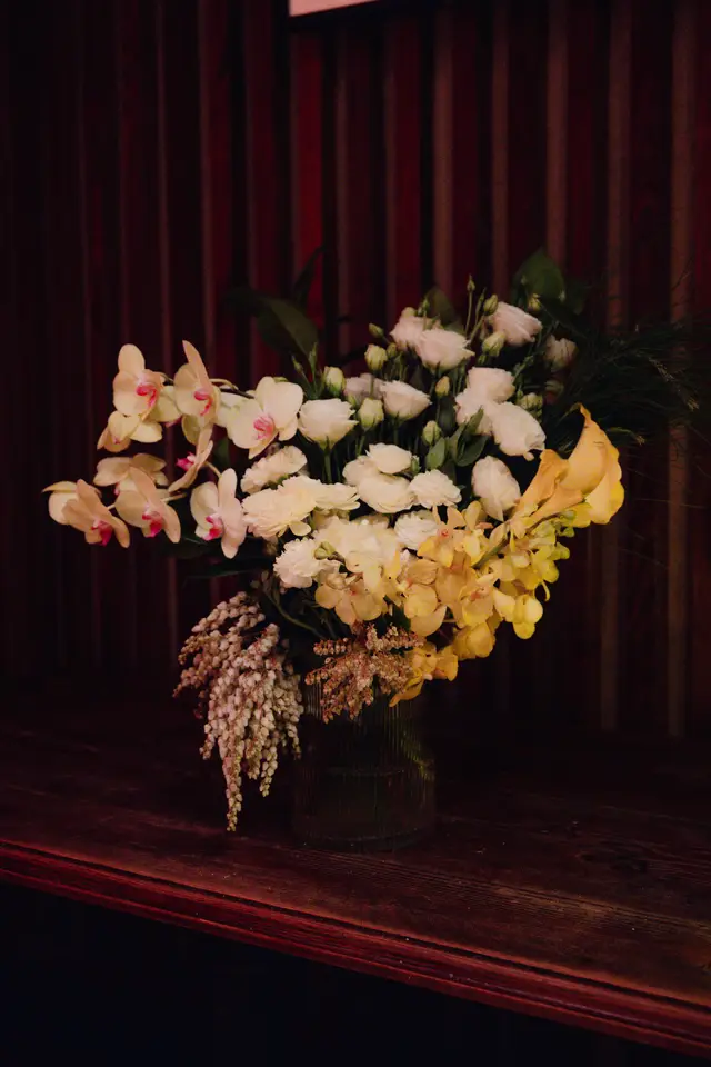 A large, elegant floral arrangement is prominently displayed in a dark glass vase on a dark wooden surface. The arrangement features a rich mix of cream, yellow, and pink flowers, with various textures and forms. The background is dark and vertically textured, contributing to a sophisticated and moody ambiance.