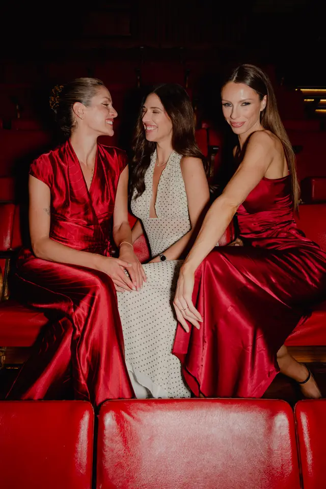 Three women are seated in red theater-style seats, smiling and interacting. Two women wear red satin-like dresses, one with a wrap style and the other a slip dress, while the third wears a white polka-dot dress. The background is dark, highlighting the subjects and the vibrant red seating.