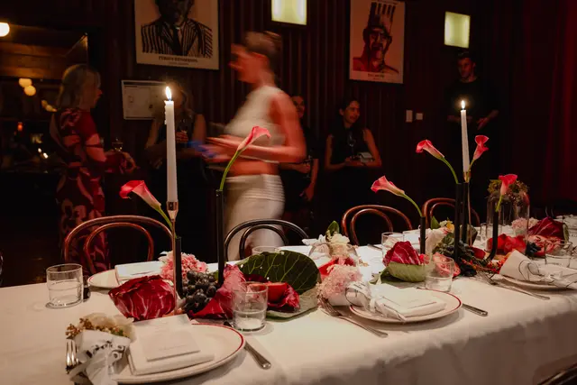 The image captures a beautifully decorated dining table at a dimly lit event. The foreground features an elaborate centerpiece with pink calla lilies, various flowers, grapes, and pomegranates, set with white plates and lit candles. In the blurred background, several guests are visible, suggesting an intimate and lively atmosphere.