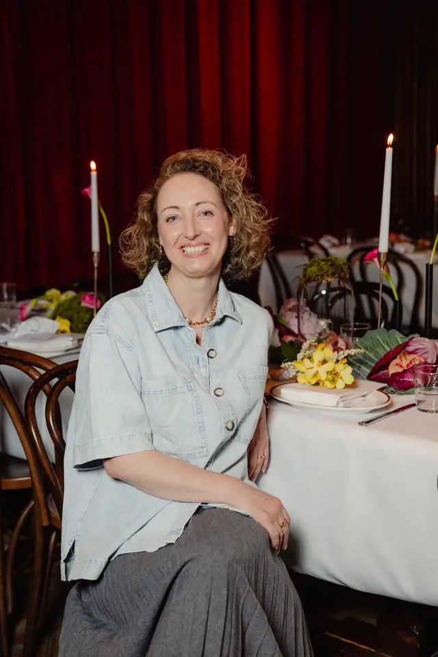 A smiling woman with curly hair, wearing a light blue denim shirt and grey pleated skirt, is seated at a formal dinner table. The table is elegantly set with a white tablecloth, lit candles, and vibrant floral arrangements featuring yellow and pink flowers, against a backdrop of deep red curtains. She is looking directly at the camera with a warm expression.