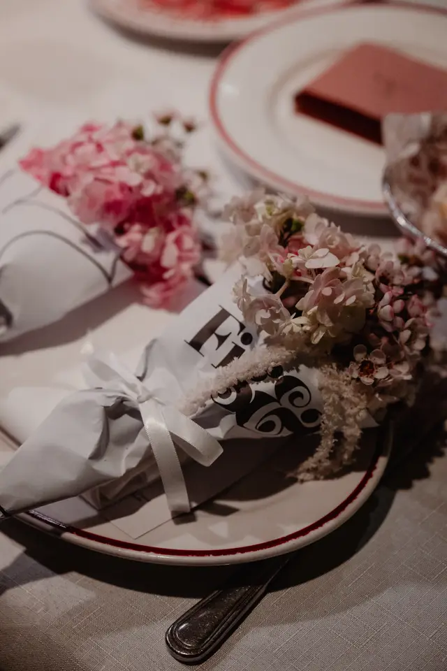 A close-up, overhead shot of two small floral bouquets wrapped in white paper, resting on white plates with red rims. The bouquets feature a mix of pink and white flowers, tied with white ribbons. In the background, a blurred dessert or pastry is visible on another plate, suggesting a formal event or dining setting.