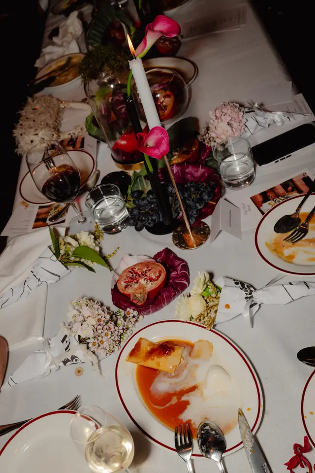 A high-angle shot captures an elegantly set table at an event, featuring partially eaten dishes, wine, water, and elaborate floral and fruit decorations. A lit white candle adorned with a pink calla lily stands as a centerpiece, surrounded by dark grapes, radicchio leaves, and a halved pomegranate. The scene conveys a sophisticated and intimate atmosphere, highlighting the detailed table styling.
