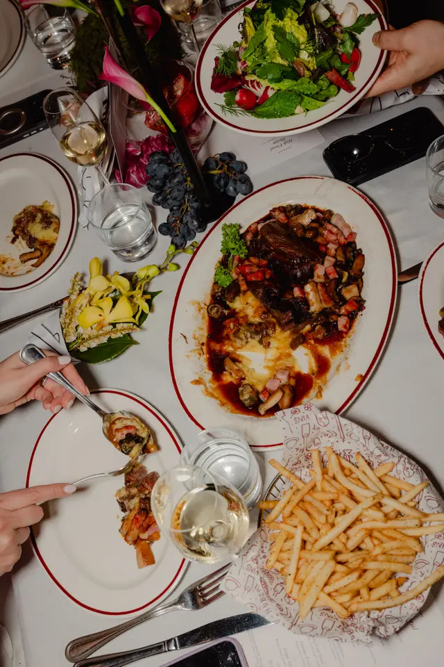 An overhead shot captures a lively dinner table scene, featuring various dishes like a hearty meat entree, fresh salad, and french fries, alongside wine and water glasses. Multiple hands are visible, engaged in dining, with elegant floral arrangements and grapes adorning the white tablecloth.