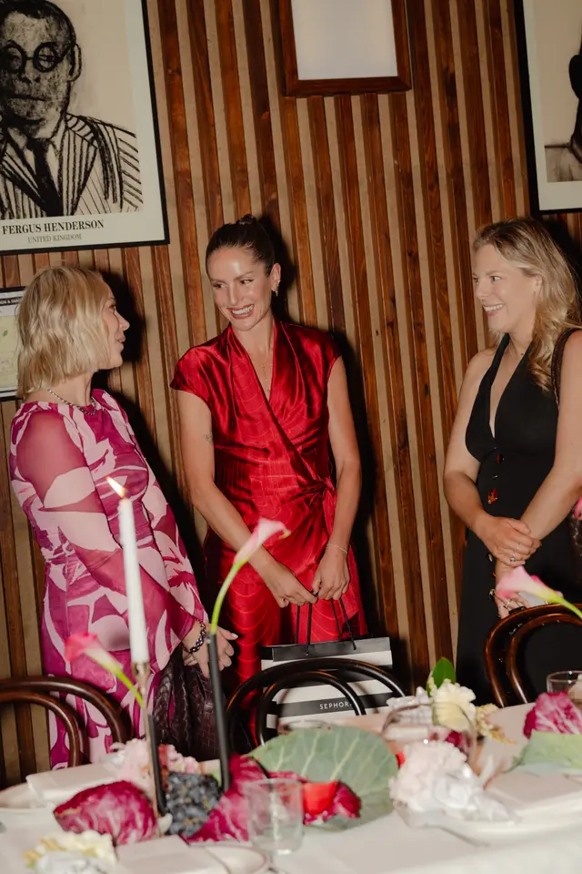 Three women are captured mid-conversation and smiling at what appears to be a formal event or dinner. They are standing around a table adorned with floral arrangements, a lit candle, and place settings. The background features a warm wooden paneled wall with framed artwork.