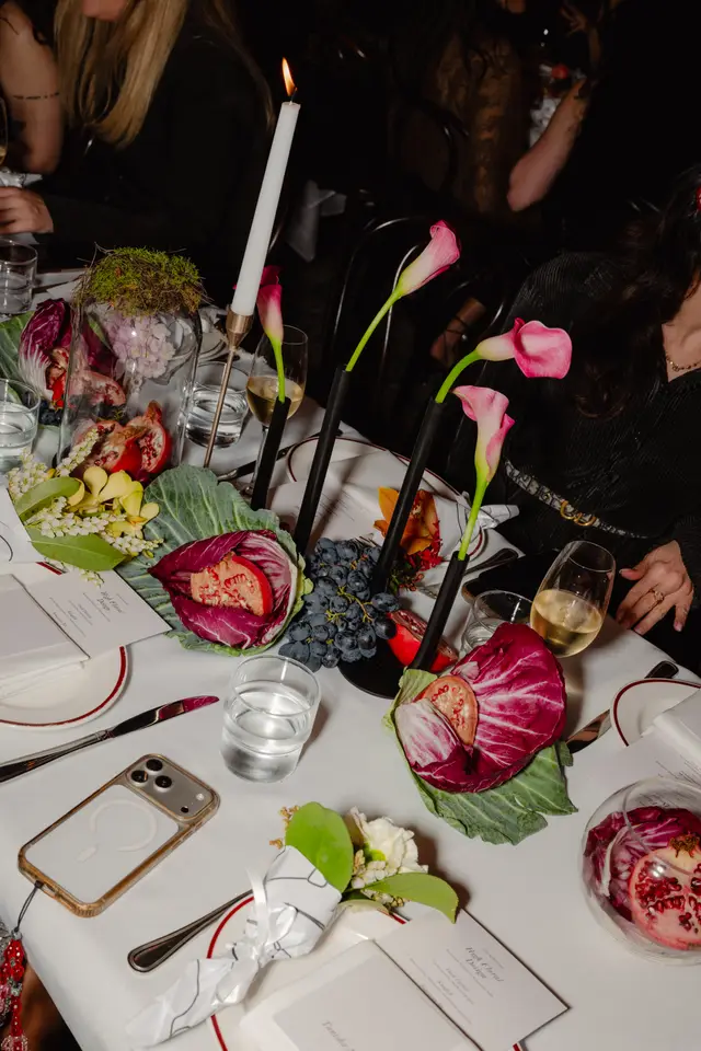 A close-up, overhead shot of an elegantly set dinner table at an event, featuring a white tablecloth. The table is adorned with various floral arrangements, fresh fruits like halved pomegranates and dark grapes, and individual place settings with menus. Lit candles and modern black vases with pink calla lilies contribute to the sophisticated ambiance.
