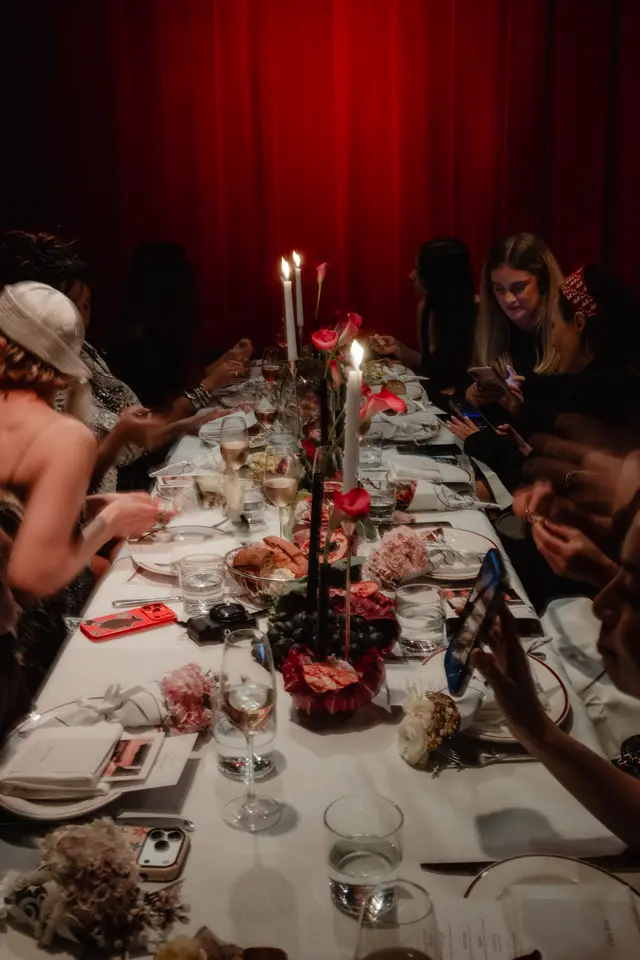A long dining table is set for an event, illuminated by candlelight and featuring elaborate floral arrangements. Multiple people are seated around the table, some interacting with their phones, against a backdrop of rich red curtains. The atmosphere appears intimate and festive.