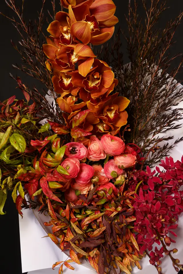 A vibrant, close-up studio shot of a rich floral arrangement. It features a prominent cascade of deep orange orchids, clusters of pink ranunculus, and small red flowers, all nestled among various green, red, and brown foliage. The arrangement is presented against a dark, almost black background, with some white wrapping paper visible at the bottom, highlighting the diverse textures and warm color palette of the blooms.