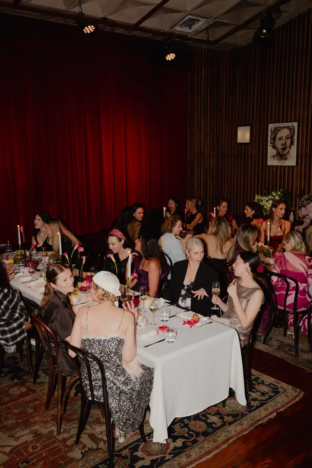 A vibrant indoor event scene captures multiple women seated around elegantly set dining tables. The room features rich red curtains, dark wooden paneling, and a patterned rug, creating a sophisticated atmosphere. Tables are adorned with white tablecloths, candles, wine glasses, and various floral arrangements, including pink calla lilies.