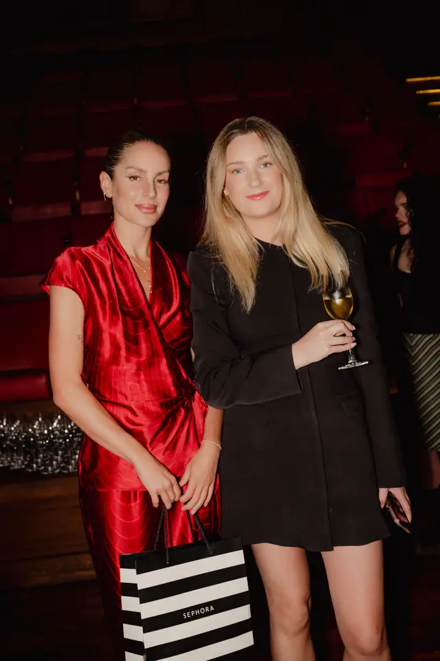 Two women pose for a photo at an indoor event. The woman on the left wears a red satin dress and holds a black and white striped Sephora shopping bag. The woman on the right wears a black blazer dress and holds a glass of champagne. They are positioned in front of dimly lit red seating, likely in a theater or event venue.