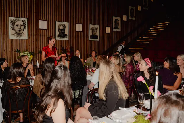 A lively indoor scene captures a group of people seated around dinner tables at an event. A woman in a red top stands and appears to be speaking to the guests, who are engaged and smiling. The setting is an upscale restaurant with dark wood paneling, framed portraits of chefs, and a grand staircase in the background.