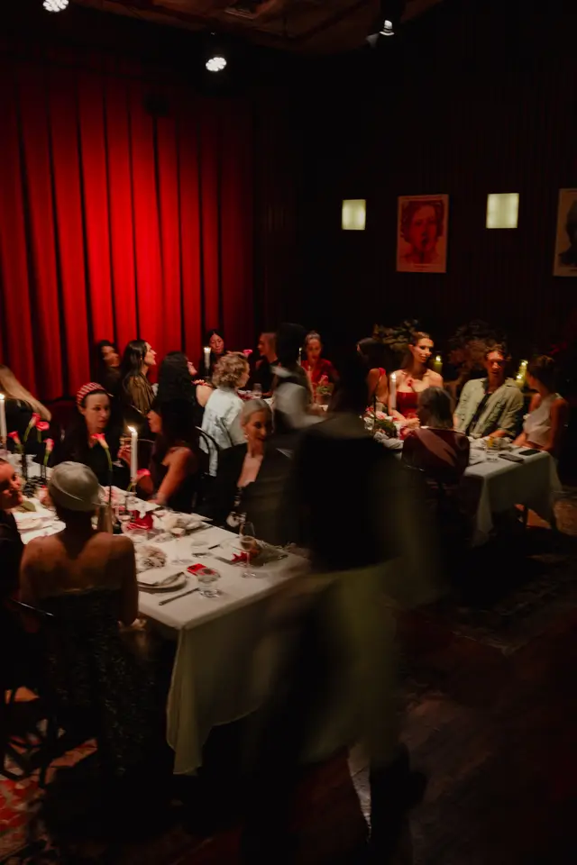A dimly lit indoor event shows multiple people seated at dining tables covered with white tablecloths. Candles and red tulips are visible on the tables, creating a warm ambiance. A large red curtain serves as a backdrop, and some figures are blurred due to motion, suggesting activity.