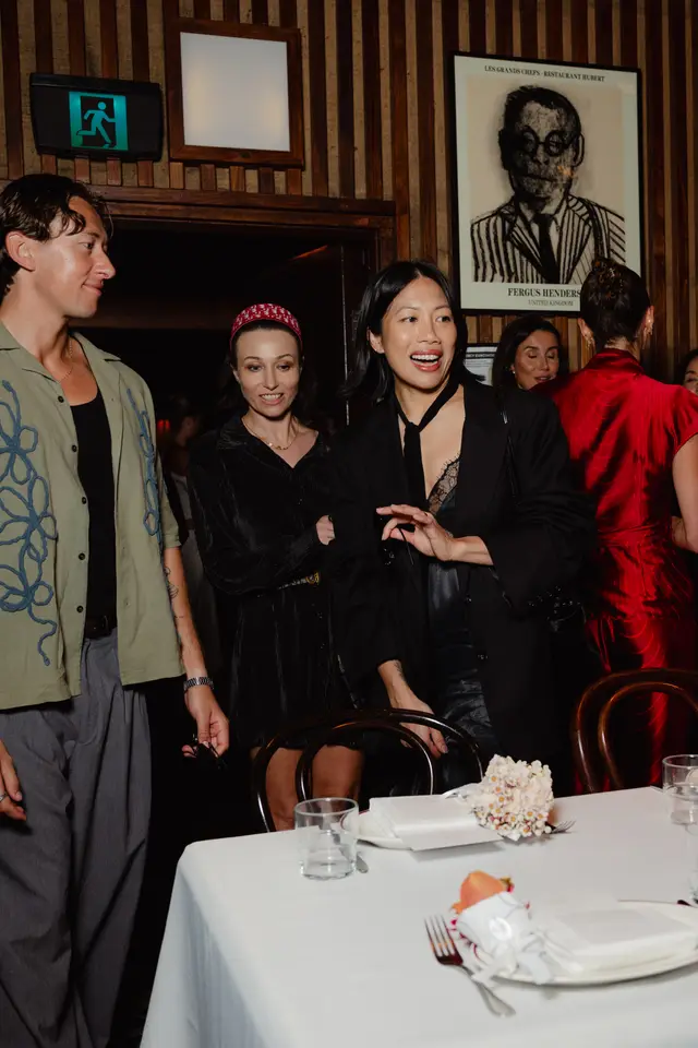 A lively indoor event scene captures several well-dressed individuals mingling around a set dining table. The atmosphere is warm and intimate, characterized by dark wood paneling and subtle lighting. A small, delicate floral arrangement serves as a centerpiece on the white-clothed table.