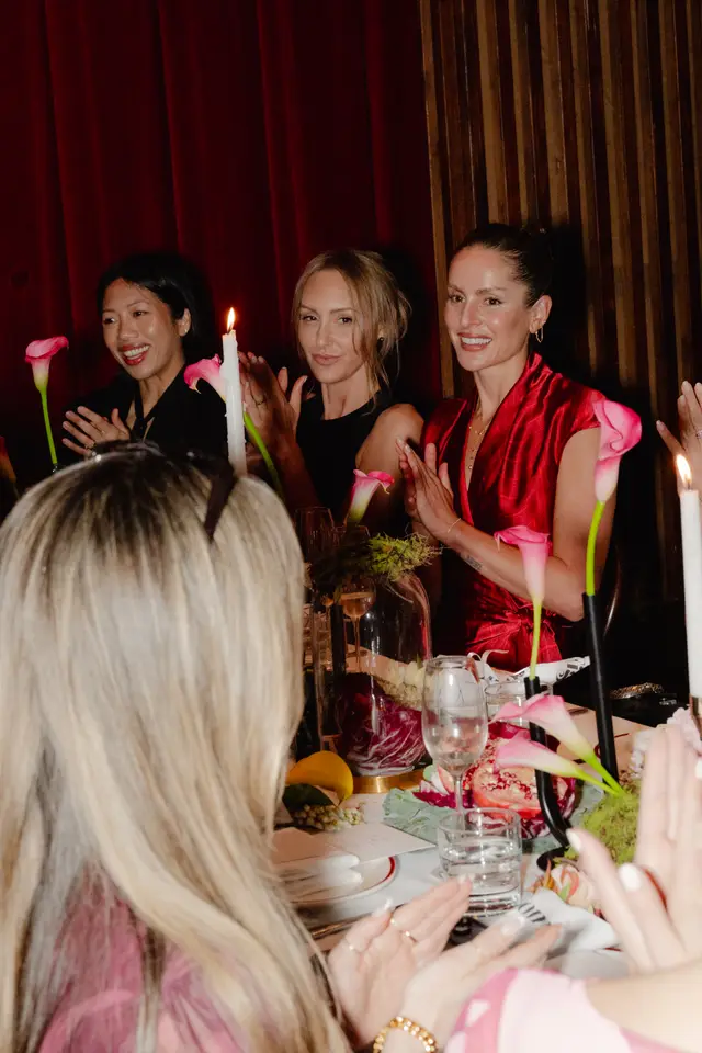 A warm, intimate shot of multiple women clapping and smiling at a dinner table, adorned with pink calla lilies, lit candles, and various fruits. The setting features deep red curtains and elegant table decor, suggesting a celebratory event or dinner party. The foreground shows the back of a person's head, also clapping.