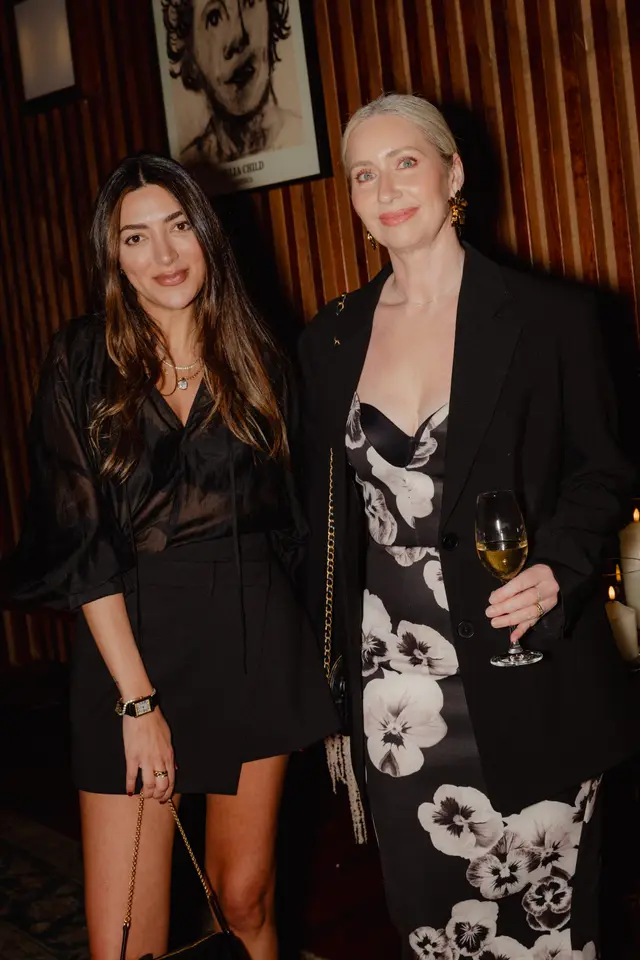 Two women are captured in a warm, dimly lit indoor setting, likely at an evening event. The woman on the left wears a black sheer top and skirt, accessorized with layered gold necklaces and a watch. The woman on the right wears a black blazer over a black and white floral dress, holding a glass of white wine.