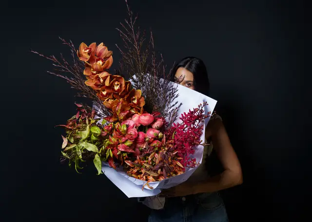 A large, vibrant floral bouquet, wrapped in white paper, is held by a person whose face is partially obscured by the flowers. The bouquet features prominent orange orchids, pink ranunculus, and various red and green foliage against a plain dark background. The person's eyes and dark hair are visible above the bouquet.