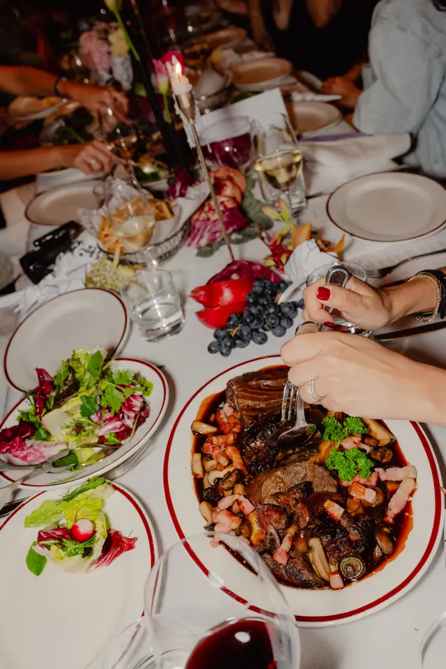 A high-angle shot captures a lively dinner table scene, focusing on a large plate of braised meat with mushrooms and bacon being served. Multiple hands are visible around the white tablecloth, holding cutlery and glasses of wine and water. Various side dishes, including fresh salads and decorative elements like grapes and peppers, are also arranged on the table, illuminated by a flickering candle.
