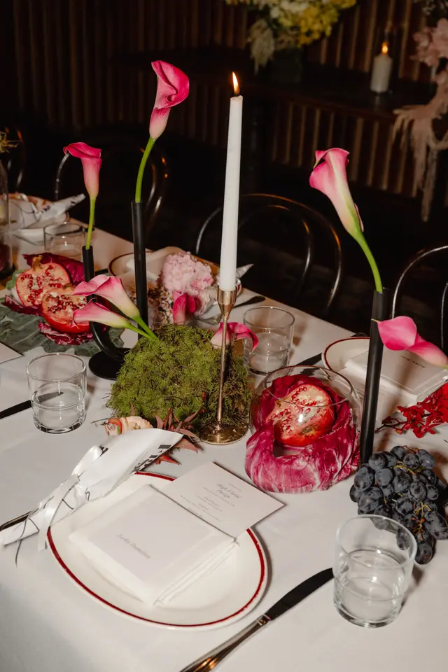 A beautifully arranged table setting for an event, featuring vibrant pink calla lilies in sleek black vases, alongside fresh pomegranates, dark grapes, and other floral and natural elements. A lit white candle in a gold holder adds a warm glow, highlighting the elegant place settings and creating an inviting atmosphere.