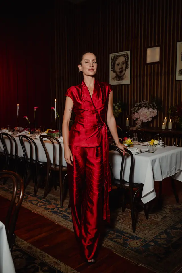 A woman in a striking red patterned wrap top and matching wide-leg pants stands beside a long dining table set for an event. The table is adorned with white tablecloths, lit candles, and various floral arrangements. The setting features dark wood paneling, framed portraits, and a patterned rug, creating an elegant and intimate atmosphere.