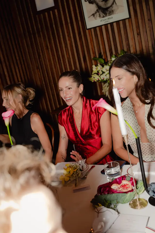 A vibrant indoor shot captures three women smiling and interacting at a well-decorated dinner table. The setting features warm lighting, a slatted wooden wall, and elegant floral and fruit arrangements including pink calla lilies, yellow orchids, pomegranate, and grapes. The atmosphere appears social and celebratory, with a framed 'Julia Child' print visible on the wall.
