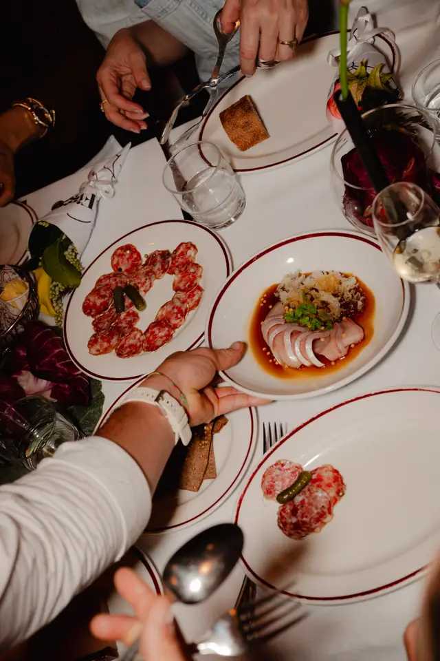 A high-angle shot captures a lively dining scene with multiple hands interacting around a white-clothed table. Various dishes are presented, including plates of sliced salami with pickles, a dish of thinly sliced fish with sauce and garnish, and bread. The setting suggests a social and elegant gathering.