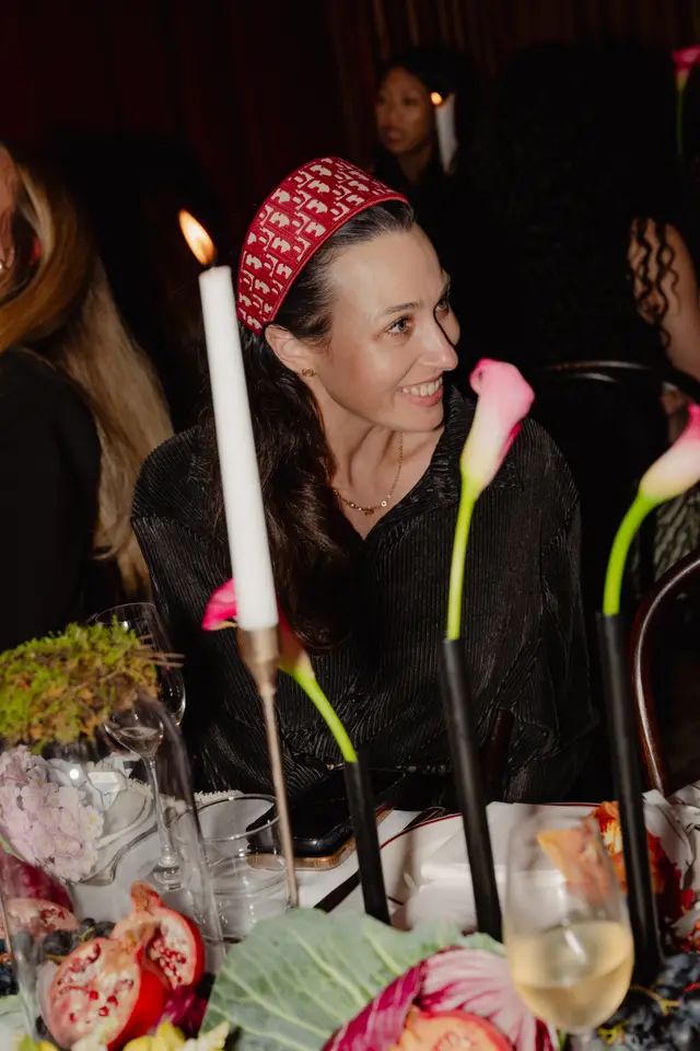 A close-up shot captures a smiling woman wearing a red patterned headband and a dark pleated shirt, seated at a dimly lit table. The table is elegantly set with a lit candle, wine glasses, pink calla lilies, pomegranates, grapes, and various greenery. Other guests are partially visible in the soft-focus background, contributing to a festive atmosphere.