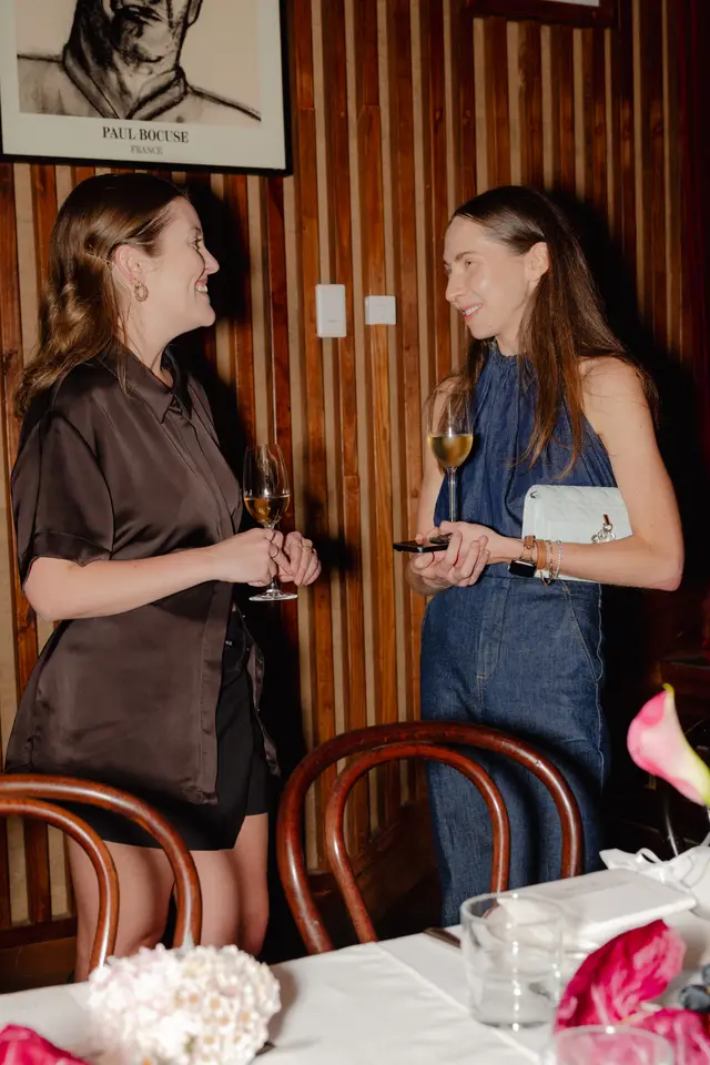 Two women are engaged in conversation at an indoor event, each holding a glass of white wine. They are positioned near a table with a white tablecloth and floral arrangements, against a warm wooden slatted wall featuring a framed portrait of Paul Bocuse.