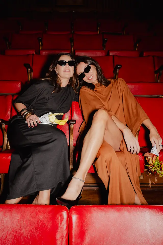Two women wearing sunglasses are seated in a row of red theater seats, looking directly at the camera. The woman on the left wears a dark grey dress and holds a small white and yellow floral arrangement, while the woman on the right wears a rust-colored dress and holds a small bouquet of green and orange foliage. The background shows more rows of empty red seats, dimly lit.