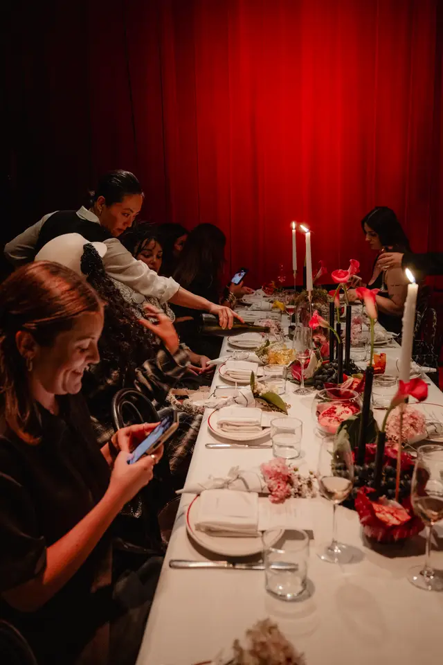 A warm, intimate scene captures multiple people gathered around a long dining table, richly decorated with floral arrangements, lit candles, and fresh fruit. Guests are seen interacting, pouring wine, and using smartphones, set against a dramatic red curtain backdrop. The focus is on the elegant table setting and the lively atmosphere of the event.