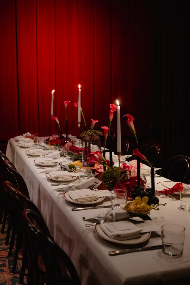 A long dining table is elegantly set for an event, featuring multiple place settings with white plates, folded napkins, and cutlery. The table is adorned with elaborate floral arrangements, tall lit candles, and decorative elements like pomegranates and grapes. A rich red curtain serves as a dramatic backdrop, creating a warm and intimate atmosphere.