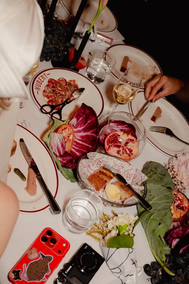 An overhead shot captures a vibrant dinner table setting, featuring an array of dishes including cured meats, pate, bread, pomegranates, and grapes, alongside glasses of wine and water. The table is adorned with floral elements and large green leaves, while personal items like a smartphone and camera are also visible, suggesting a lively event.