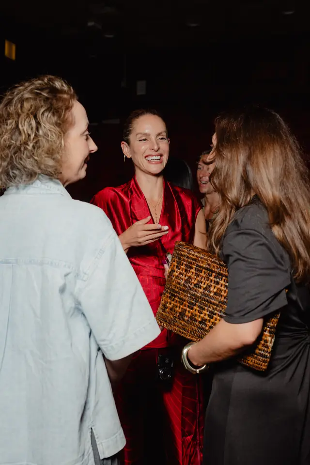 A candid, medium shot captures multiple women interacting and laughing at an indoor event. The central figure, wearing a red patterned top and gold jewelry, is laughing heartily, with her hand raised to her chest. Other women are partially visible around her, one in a denim shirt and another holding a woven clutch.