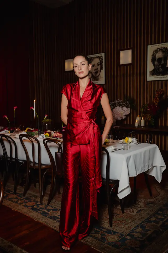 A woman in a striking red patterned wrap top and matching wide-leg pants stands in a dimly lit room. Behind her, a long dining table is set with a white tablecloth, candles, and floral arrangements. The background features dark wood paneling and framed artwork, suggesting an elegant event setting.