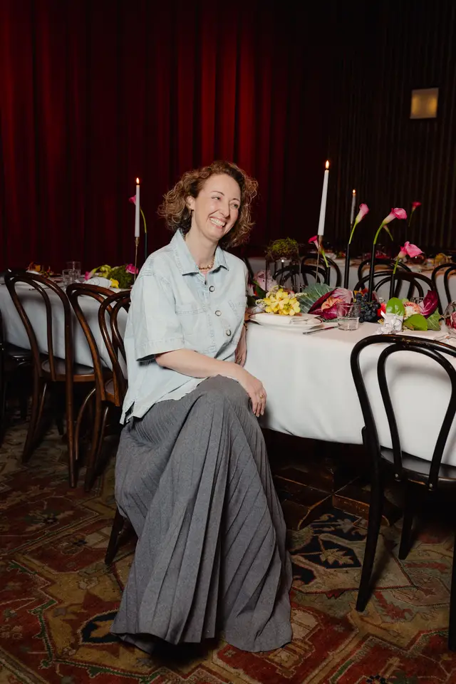 A smiling woman with curly hair is seated at a beautifully decorated event table, looking towards the right. She is wearing a light blue denim shirt and a long grey pleated skirt. The table is adorned with white tablecloths, candles, various flowers, fruits, and vegetables, set against a backdrop of dark red curtains.