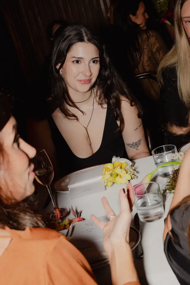 A candid, close-up shot captures a woman with dark hair and a black top, smiling subtly while seated at a dimly lit table. She wears a distinctive silver necklace and has a tattoo on her arm. The table is elegantly set with floral arrangements featuring yellow orchids and pink calla lilies, along with menu cards and drinks, suggesting a sophisticated social event.
