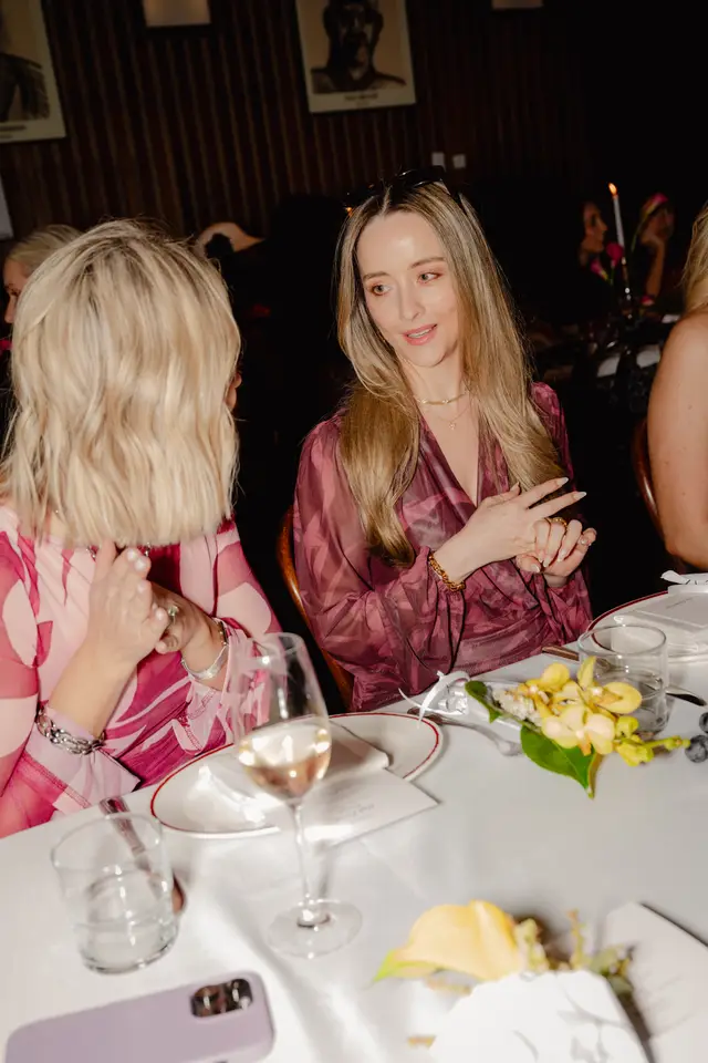 A vertical shot captures two women seated at a table during an indoor event. The woman on the right, with long blonde hair, is actively speaking and gesturing, while the woman on the left, with shorter blonde hair, is listening. The table is set with white tablecloths, plates, glasses, and a small arrangement of yellow flowers.