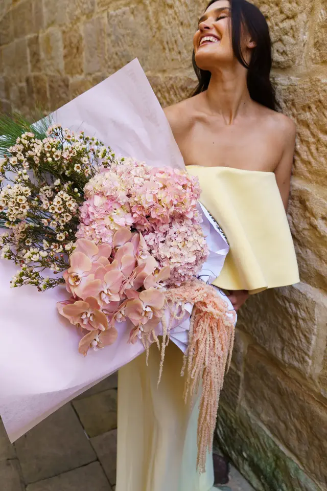 A smiling woman in a light yellow strapless dress holds a large, vibrant bouquet of pink, peach, and white flowers. She is looking upwards with a joyful expression, leaning against a textured stone wall. The bouquet features hydrangeas, orchids, waxflowers, and cascading amaranthus, wrapped in pink and white paper.